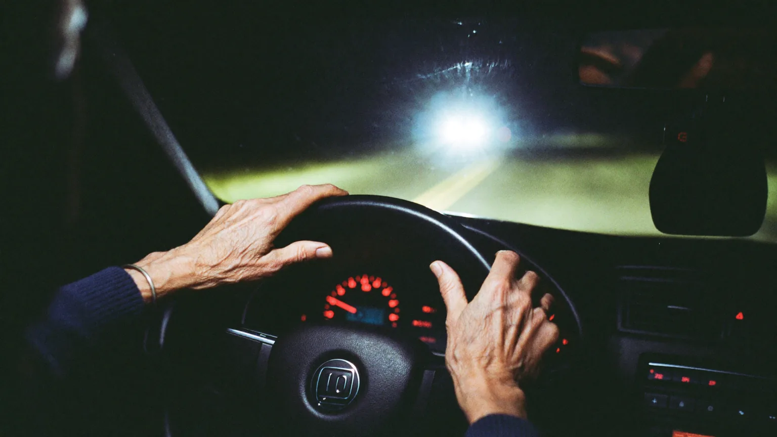Older driver's hands on a steering wheel at night with oncoming headlights producing glare halos