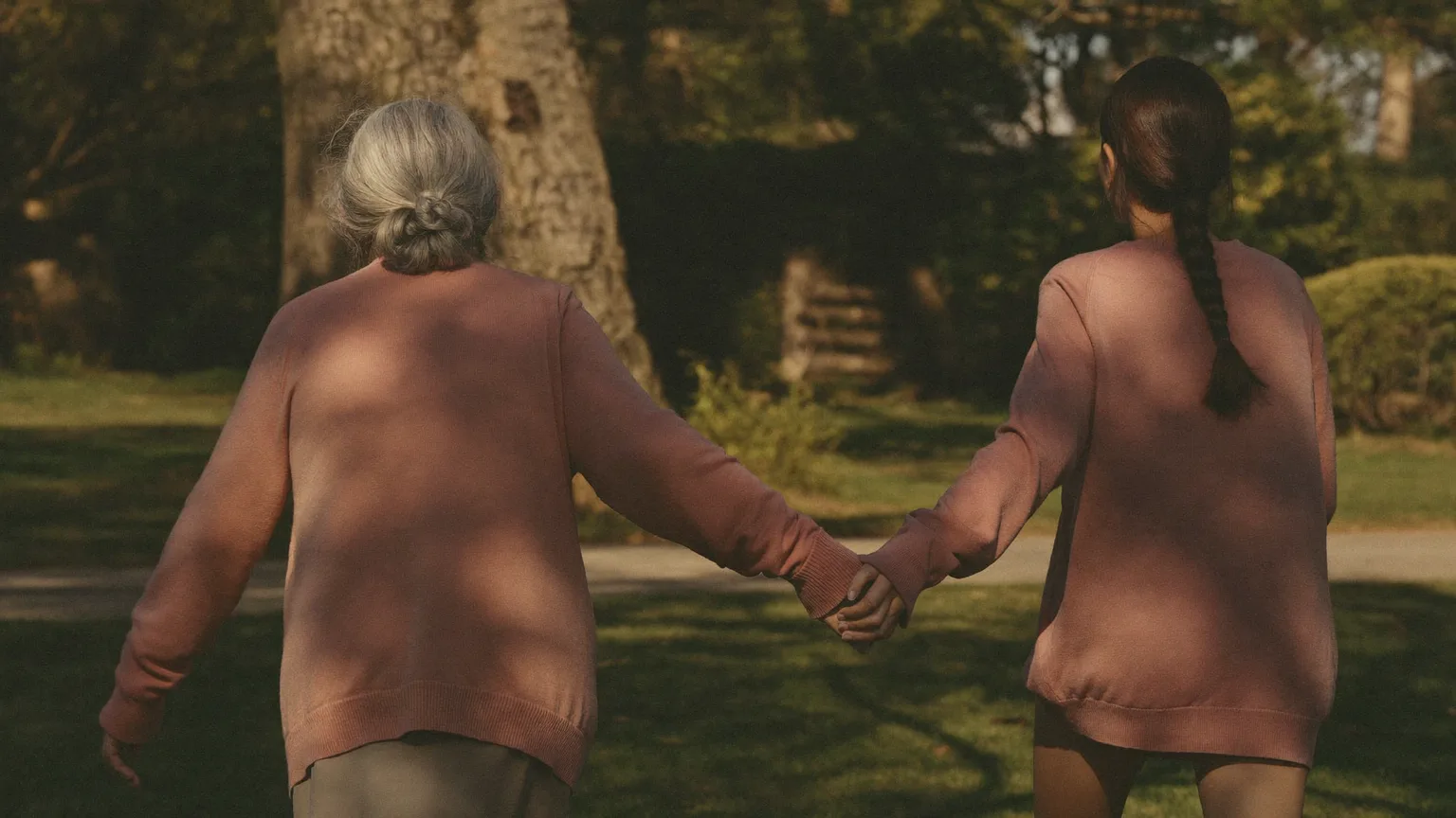 Elderly mother and her adult daughter walking arm in arm along a sunlit garden path