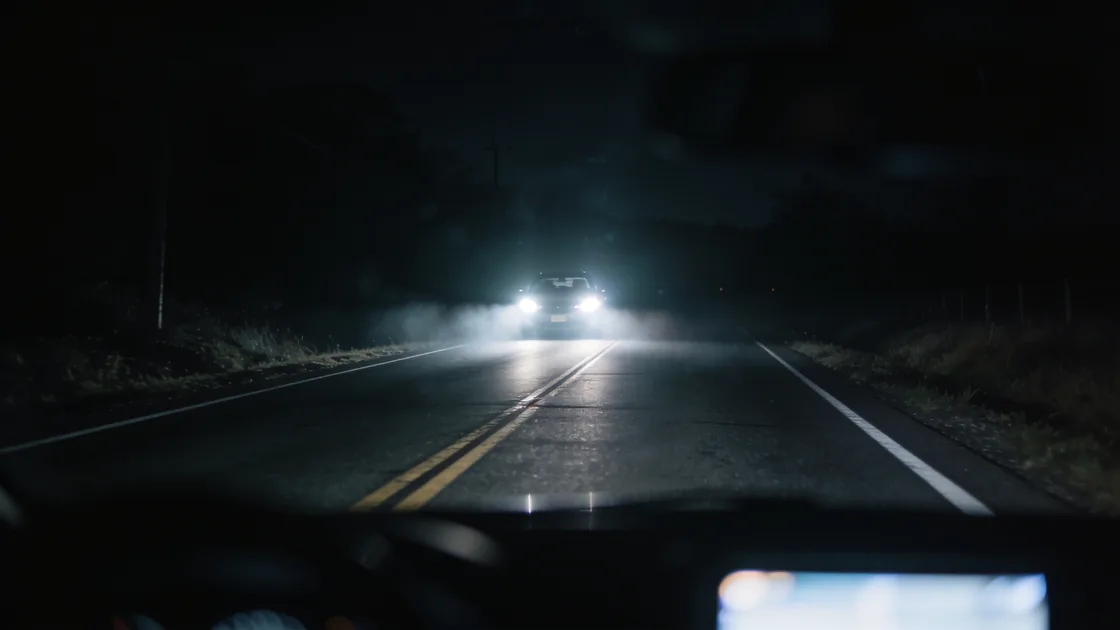 Oncoming headlights flaring on a dark rural road seen from the driver's seat