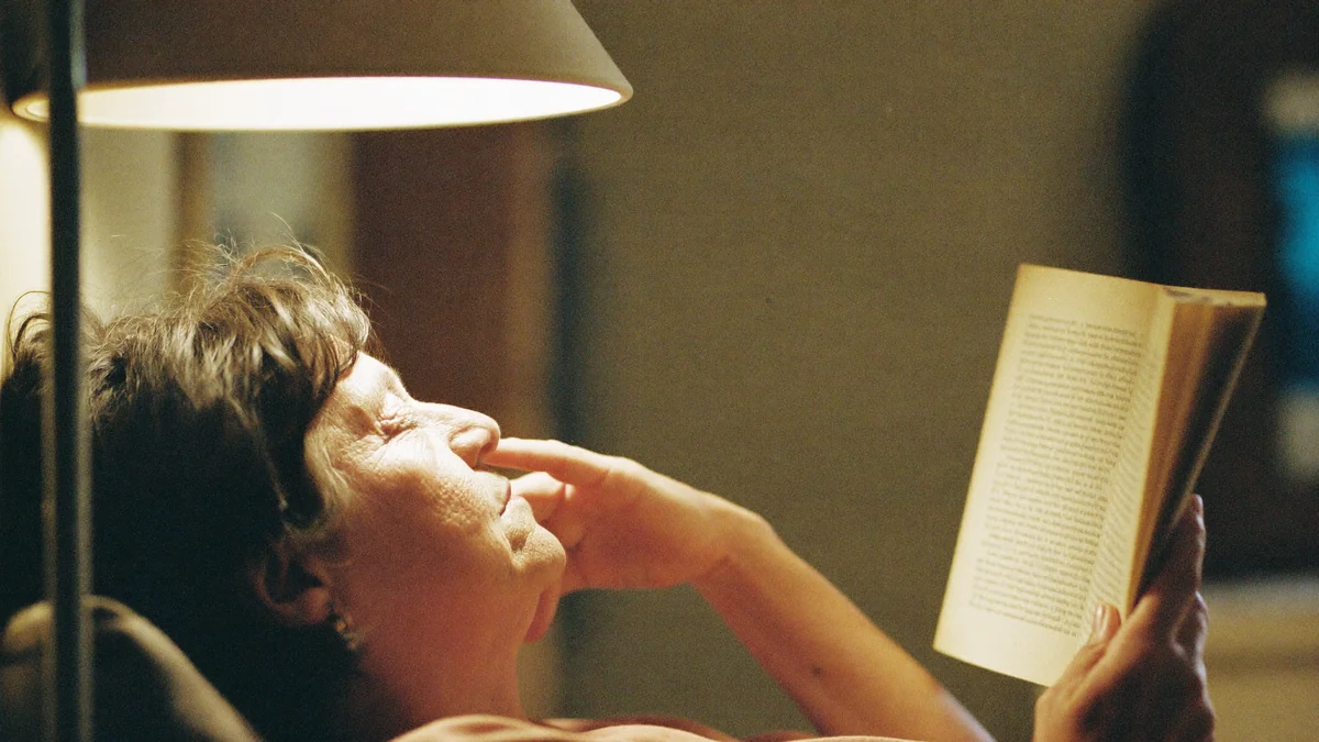 Older woman leaning back from a book, pressing the bridge of her nose with eye fatigue